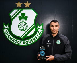 A man holding a soccer ball in front of a green and white logo.